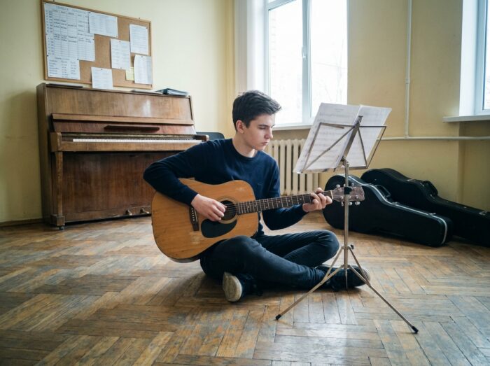 Adolescent jouant de la guitare acoustique seul dans une salle de répétition de conservatoire, partitions devant lui, ambiance studieuse